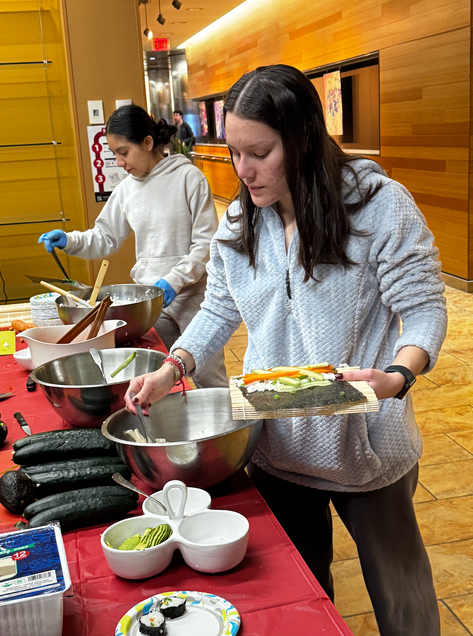  On the right a student is adding tofu to her nori paper, that already has rice, carrots, cucumbers, and scallions. The student on the left is shown grabbing nori seaweed paper.